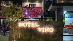 A stone sign with "HOTEL VINO" illuminated in gold letters, set against a modern building facade with a red canopy structure above. - Hotel Vino (Durrës)
