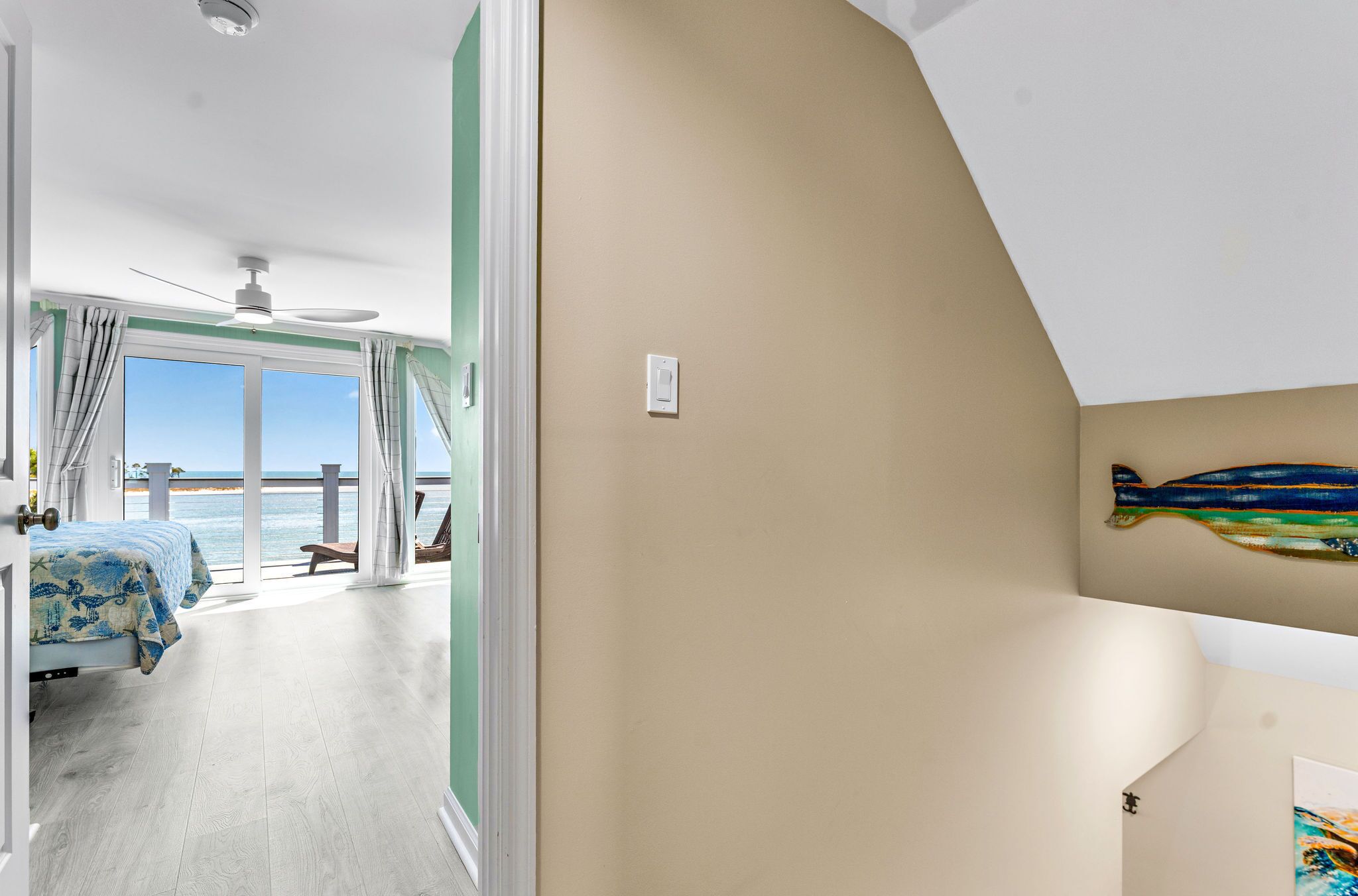 A bedroom with a bed, a ceiling fan, a view of the sea, and a wall-mounted artwork.