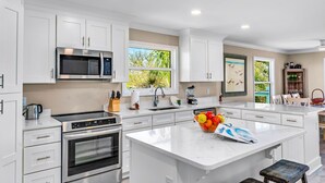 A modern kitchen with white cabinetry, stainless steel appliances, a large window, and a central island with a fruit bowl.