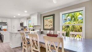 A modern kitchen with white cabinetry, a dining area with a large table and chairs, and a view of palm trees through a window.