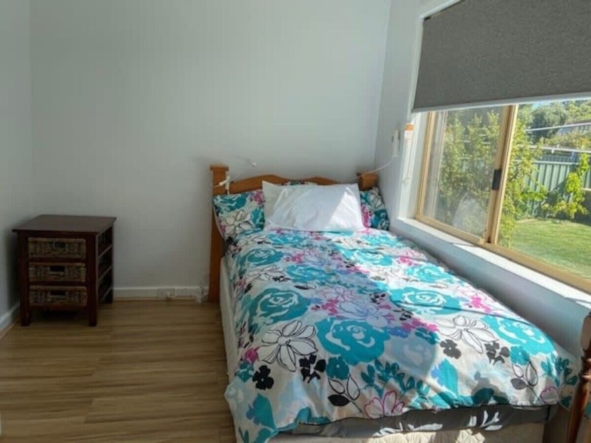 A bedroom with a wooden bed, a small wooden nightstand, and a window with a view of greenery.