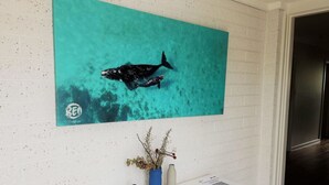 A room with a white brick wall, a large teal artwork of dolphins, a white console table with vases, and a door to the right. - Blue Tides Holiday Accommodation (Castletown)