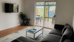 A living room with a flat-screen TV mounted on a white brick wall, a grey sofa, a white coffee table with a book and remote, and a sliding glass door leading to an outdoor patio with a table and chairs. - Blue Tides Holiday Accommodation (Castletown)