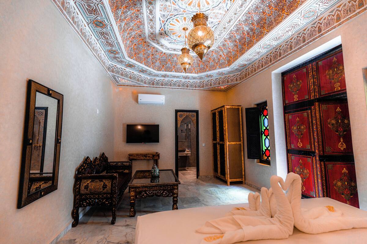 A room with a bed, a television, a mirror, a table, and a decorative ceiling.