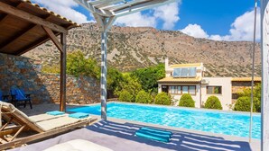 A poolside area with lounge chairs, a stone wall, and a view of a mountain and a house. - Villa Elounda Plaka - Three Bedroom Villa, Sleeps 6 (Plaka)