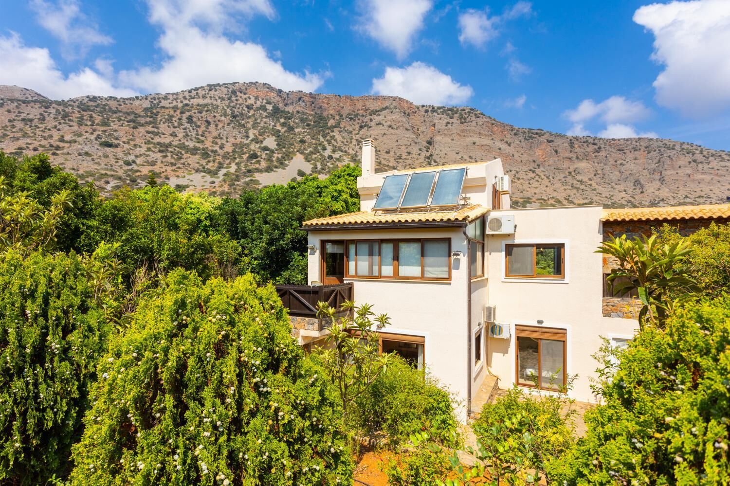 A modern two-story house with solar panels, surrounded by lush greenery and mountains in the background.