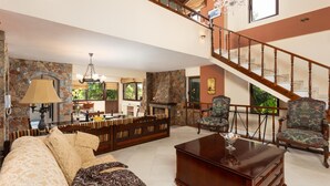Amplia sala de estar con chimenea de piedra, una escalera de madera y una zona de comedor con cojines a rayas.