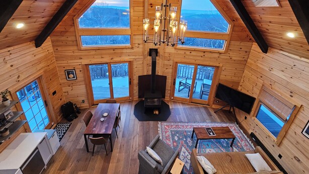 Una cabaña de madera con techos altos, chimenea central, ventanas amplias y una zona de estar acogedora.