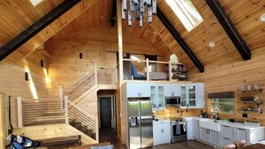 Una cabaña tipo loft espaciosa, con techos altos, una zona de cocina central, un rincón de estar acogedor con sofá y una escalera que sube a un nivel superior.