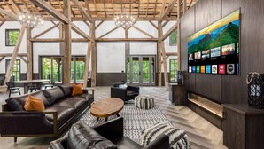 Un vaste espace de vie doté d’un haut plafond, de poutres en bois, de canapés en cuir, d’une table basse ronde et d’un grand téléviseur à écran plat fixé au mur, qui diffuse différentes chaînes.