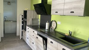 A modern kitchen with light wood cabinets, a black countertop, and a green accent wall. - Ferme Familiale Rénovée Dans un Village Tranquille (La Vendeix, GELLES)