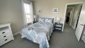 A bedroom with a bed, two bedside tables, a nightstand, a dresser, a window with curtains, and a door leading to another room.