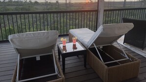 A balcony with two lounge chairs and a small table, overlooking a beach and sunset.