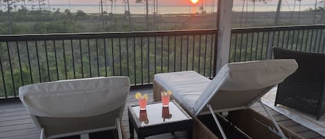 A balcony with two lounge chairs and a small table, overlooking a beach and sunset.