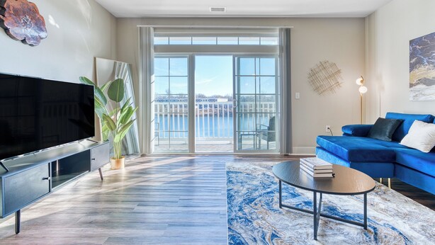 A modern living room with a large flat-screen TV, a blue sofa, a coffee table, and a view of a body of water through large sliding doors. - Ayasha River-view King Bed Suite in Keystone/Broad-Ripple/Carmel (Indianapolis)