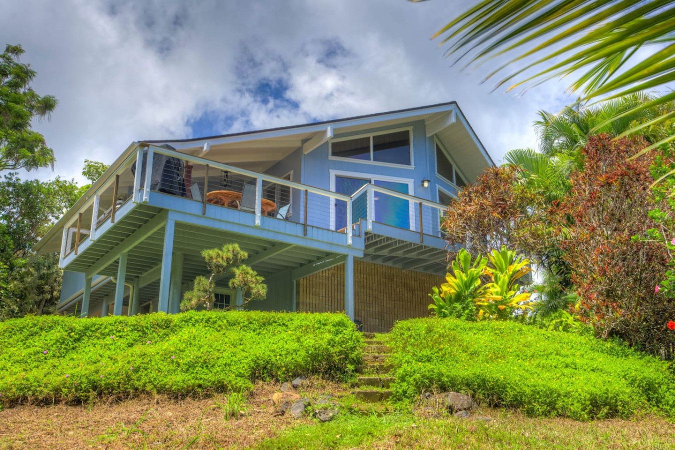 A two-story house with a balcony, surrounded by lush greenery and a clear sky.