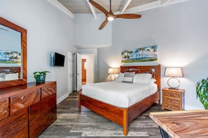 Habitación de hotel con cama de madera, ventilador de techo, televisor y un cuadro de una playa.