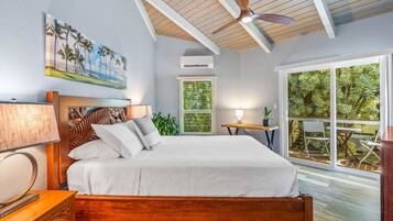 Une chambre avec un lit en bois, un ventilateur de plafond, un tableau représentant une plage et une vue sur la verdure par une grande fenêtre.