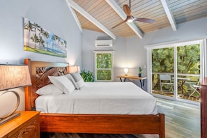 A bedroom with a wooden bed, a ceiling fan, a painting of a beach, and a view of greenery through a large window.