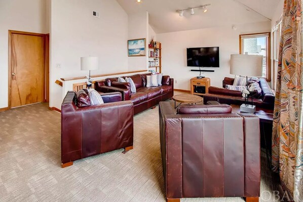 A living room with a large sectional sofa, a coffee table, and a TV. There is a bookshelf and a window with curtains. - Oceanfront Pet-Friendly House with Private Pool in Salvo (Rodanthe)