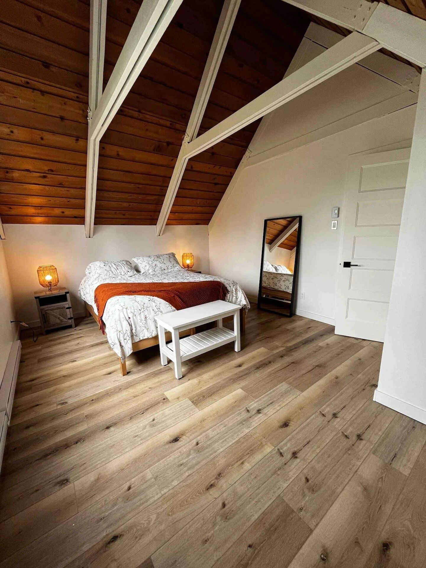 Un dormitorio con techo inclinado, piso de madera, cama con colcha blanca y naranja, mesita de noche con lámpara y espejo en la pared.