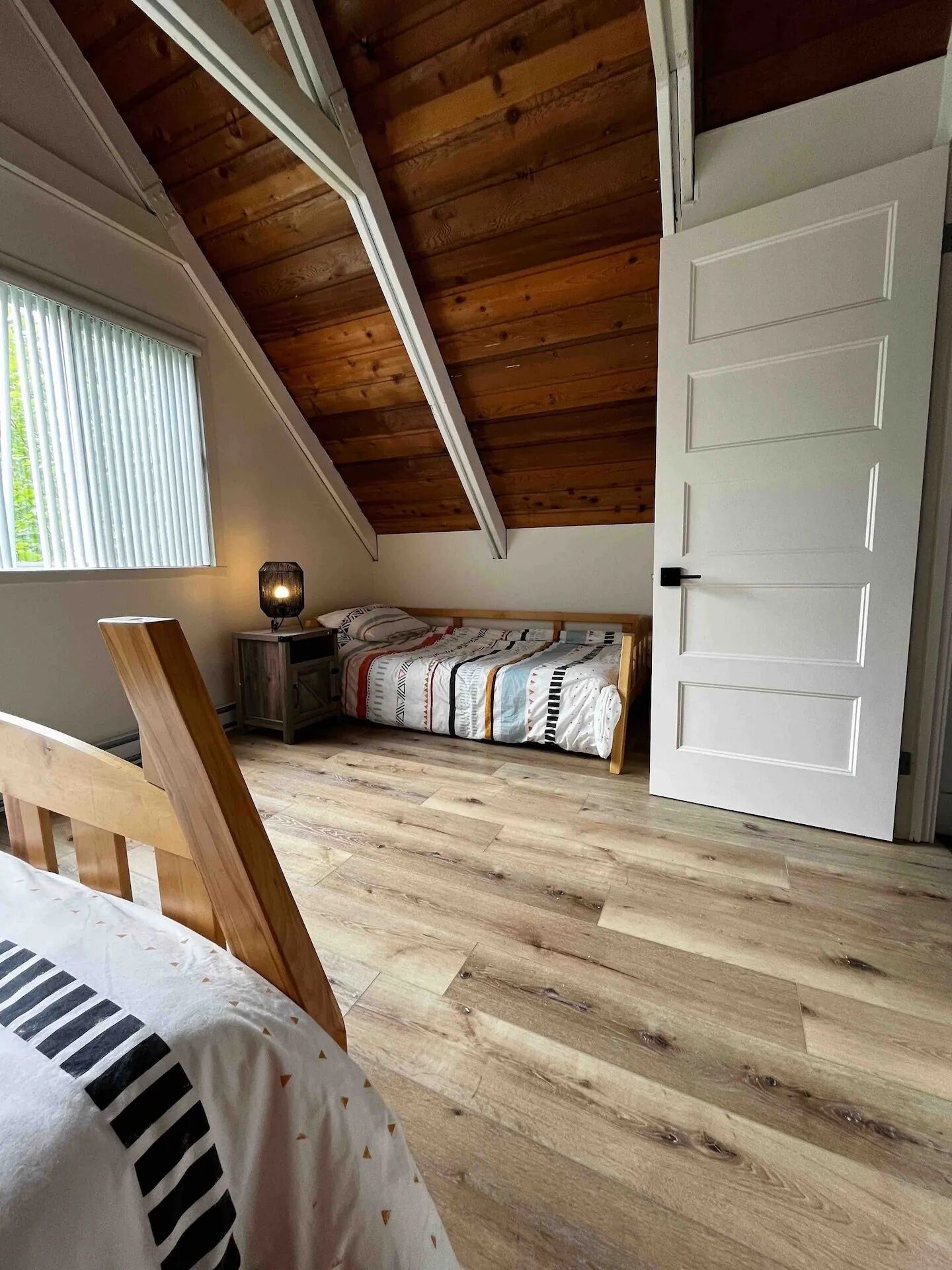 Una habitación con techo de madera inclinado, una cama con ropa de cama estampada, una mesita de noche con lámpara y una puerta.