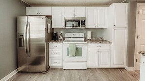 A compact kitchen with white cabinets, a stainless steel refrigerator, a microwave, and an oven. - The Bungalow on Lake Drive (Shawano)