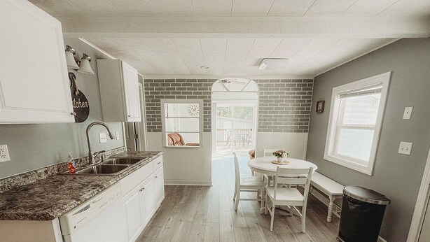 A kitchen with white cabinets, a granite countertop, a sink, and a dining area with a table and chairs. - The Bungalow on Lake Drive (Shawano)