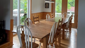 A dining area with a large wooden table and white chairs, large windows with a view of greenery, and a ceiling fan.