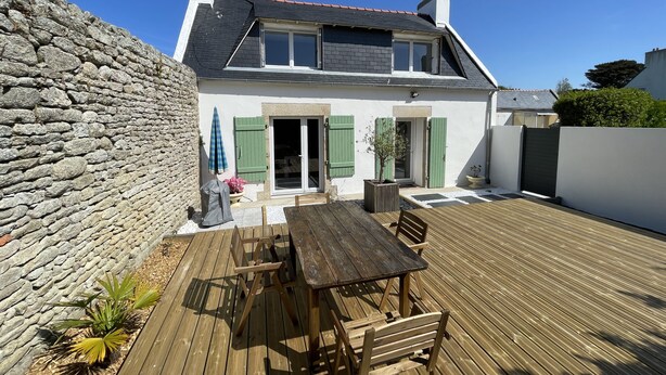 Ett hus med en stenmur, en träveranda och en uteplats med ett bord och stolar.
