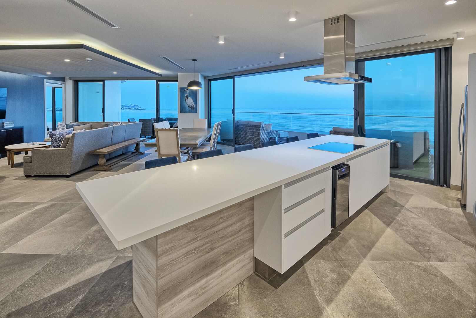 A modern kitchen with a large island, a dining area with a sofa and chairs, and a view of the sea through large windows.