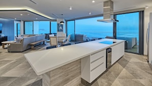 A modern kitchen with a large island, a dining area with a sofa and chairs, and a view of the sea through large windows. - LUXURY NEW OCEANFRONT CONDO- Las Palomas PH 3, 5BR/BA, 4485 Total SF , Sleeps 12 (Puerto Peñasco)