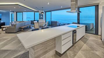 A modern kitchen with a large island, a dining area with a sofa and chairs, and a view of the sea through large windows.