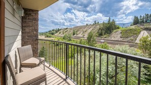A balcony with wicker furniture and a stone wall, overlooking a railway track and hilly landscape. - Waterfront Bliss | Lakeview | Pool | Hot Tub | Gym (Invermere)