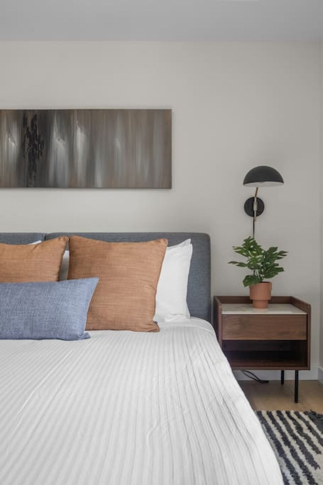 A bedroom with a bed, pillows, a nightstand, a lamp, and a potted plant.