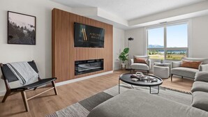 A modern living room with a fireplace, a large TV mounted on a wooden panel wall, a sofa, a coffee table, and a view of the outdoors through large windows. - Waterfront Bliss | Lakeview | Pool | Hot Tub | Gym (Invermere)