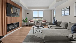 A modern living room with a flat-screen TV mounted on a wooden wall, a grey sofa, a round coffee table, and a large window offering a view of the outdoors. - Waterfront Bliss | Lakeview | Pool | Hot Tub | Gym (Invermere)