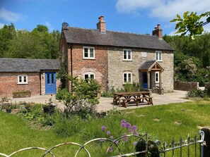 A two-story stone cottage with a blue door, a small attached shed, and a garden with purple flowers.