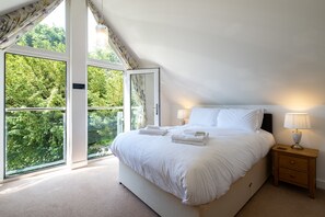 A bedroom with a large bed, two bedside tables, and a view of greenery through the windows. - Trewhiddle Villa 27 (St Austell)