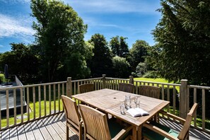 A wooden deck with a table and chairs, surrounded by a wooden fence, overlooking a grassy area and trees. - Trewhiddle Villa 27 (St Austell)