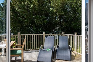A deck with two lounge chairs and a table, surrounded by a wooden railing and dense foliage. - Trewhiddle Villa 27 (St Austell)