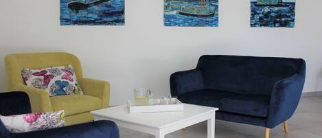 Un salón con un sillón amarillo, un sofá azul oscuro y una mesa de centro blanca. Hay tres cuadros en la pared que representan escenas marinas.