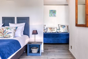 A bedroom with a bed, bedside table, lamp, and a blue sofa with patterned cushions. - L'Escapade Bellepierre (Saint-Denis)