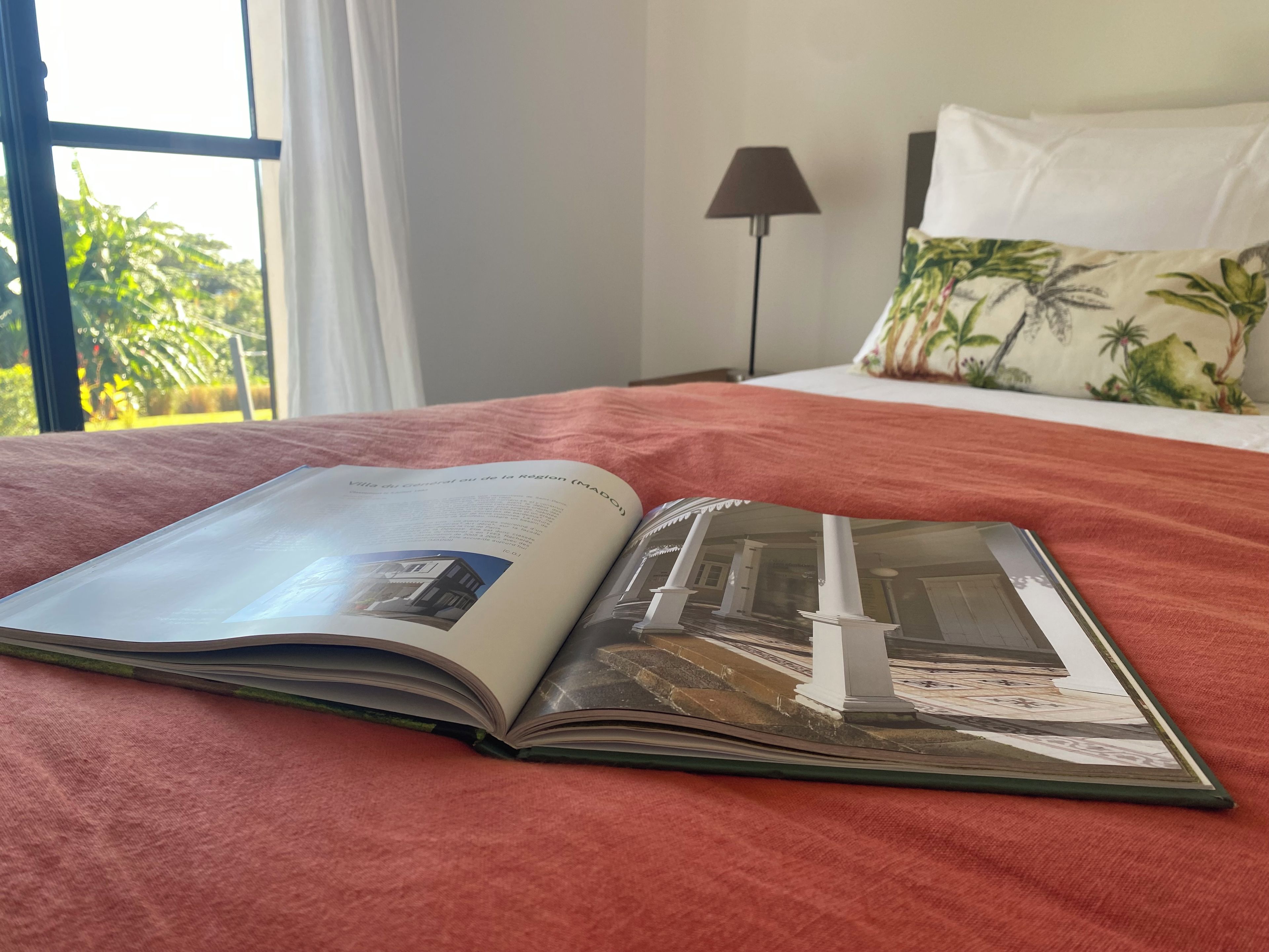 Una revista abierta sobre la cama, con una cubierta roja, una ventana con cortinas blancas y una lámpara al fondo.