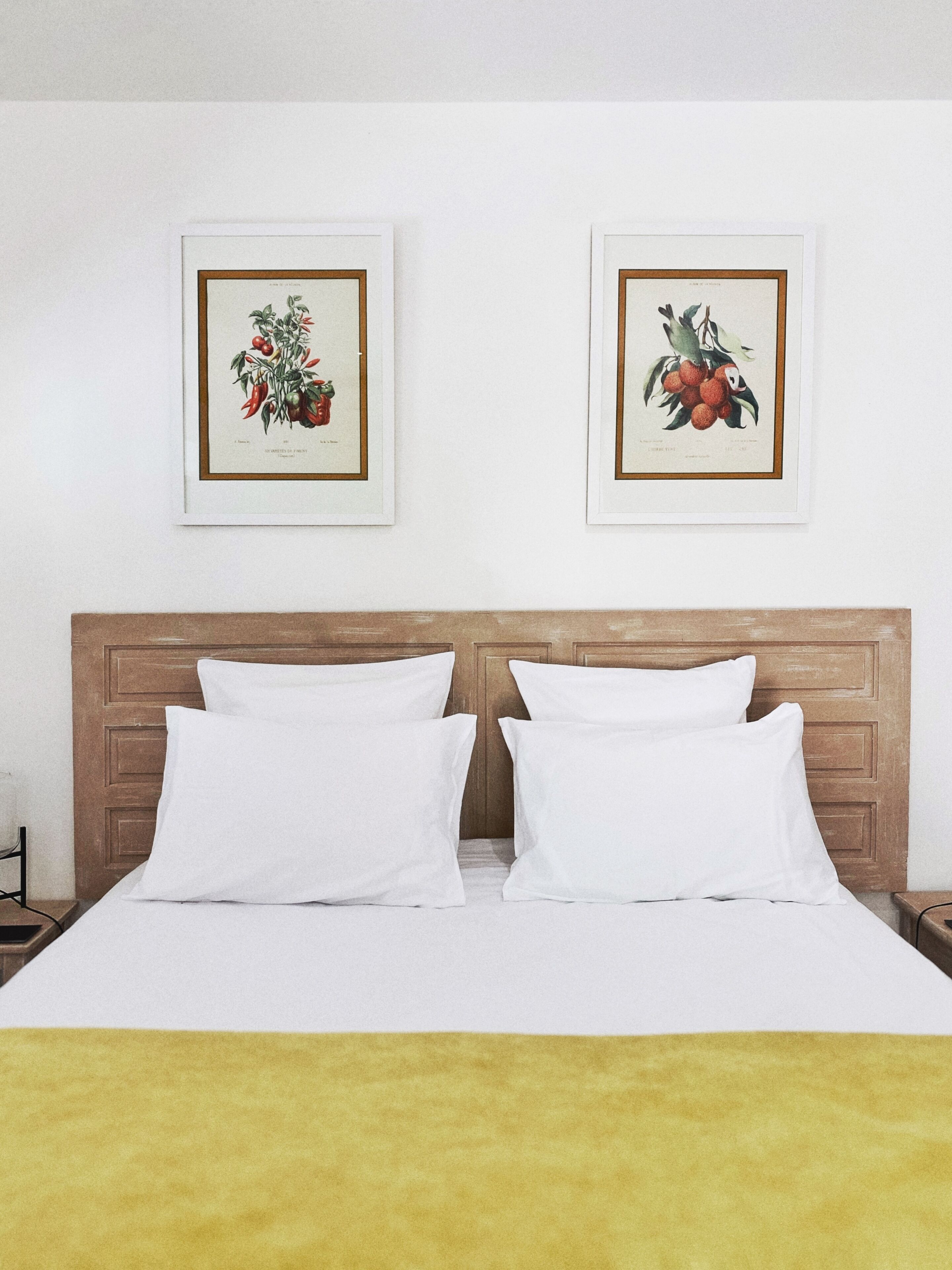 Una cama bien hecha con ropa de cama y almohadas blancas, un cabecero de madera y dos ilustraciones botánicas enmarcadas en la pared.