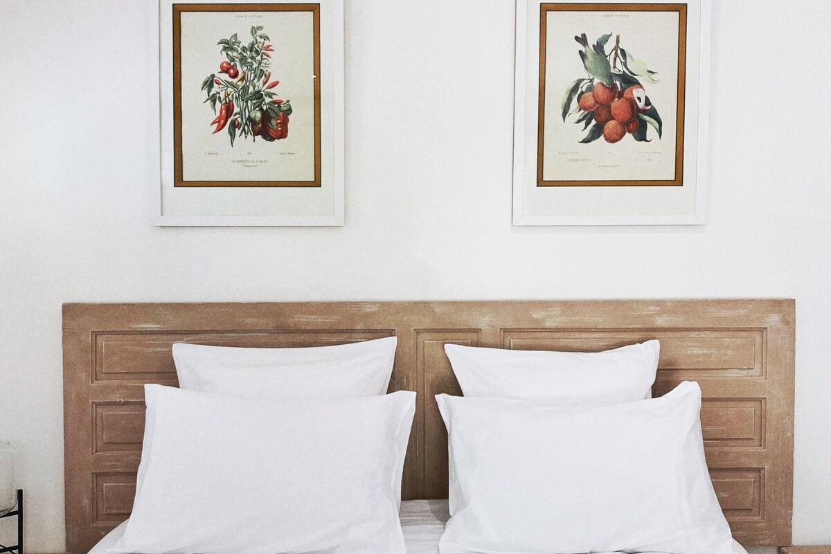 A neatly made bed with white linens and pillows, a wooden headboard, and two framed botanical illustrations on the wall.