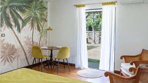 A room with a bed, two yellow chairs, a table, a wicker sofa, a mural of palm trees, and a view of a garden through a glass door. - L'Escapade Bellepierre (Saint-Denis)