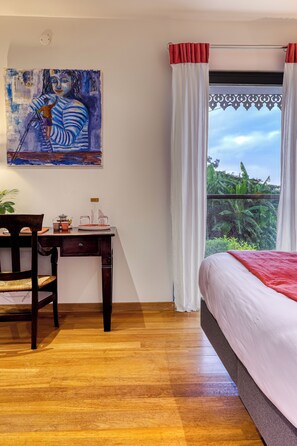 A room with a bed, a desk with a chair, a painting on the wall, and a window with curtains. - L'Escapade Bellepierre (Saint-Denis)