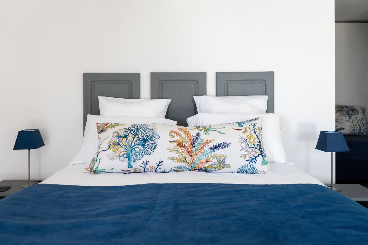 Un llit amb llençol blau, almoinos blancs i una almoina floral colorida. Hi ha dues taules de nit amb làmpades.