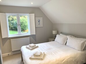 A bedroom with a large bed, white bedding, a window with shutters, a radiator, a bedside table with a lamp, and a framed picture on the wall. - Trewhiddle Villa 13 (St Austell)